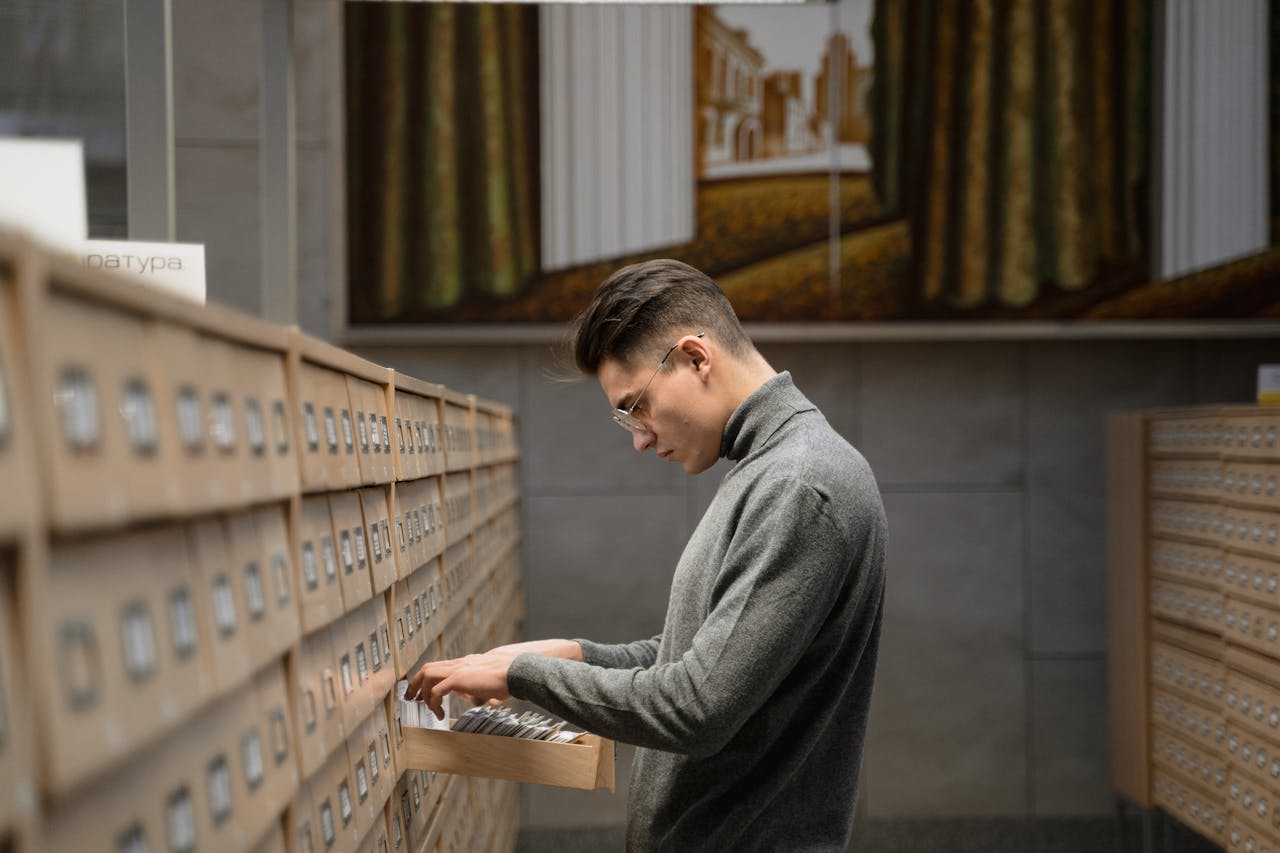 A man stands in a library looking through card files from a drawer, surrounded by bookshelves.
