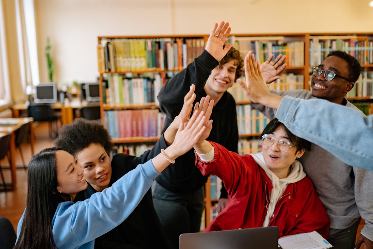 services-04 Group of happy students high-fiving in a library setting, celebrating success.